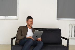 Vorderansicht eines gelangweilten jungen mannes, der auf einem sofa sitzt, während er das tablet beobachtet