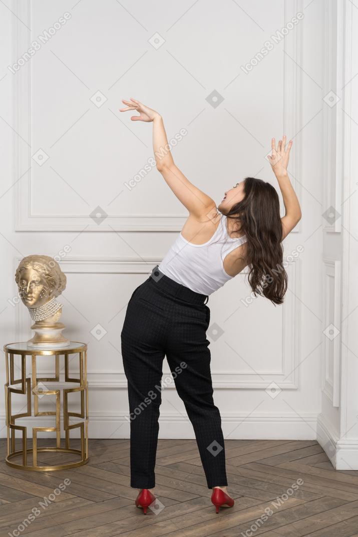 Back view of a young female raising hands and tilting body while standing near golden greek sculpture
