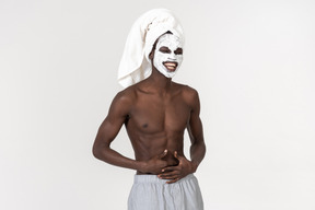 Um jovem negro de calça cinza e uma toalha na cabeça cuidando de sua rotina de cuidados com a pele