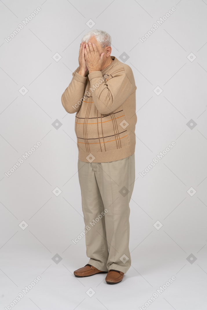 Vorderansicht eines alten mannes in freizeitkleidung, der das gesicht mit den händen bedeckt