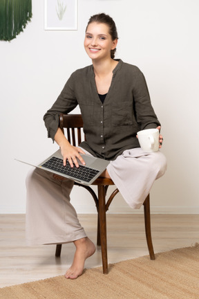 Vue de face d'une jeune femme portant des vêtements de maison assise sur une chaise avec un ordinateur portable et buvant du café