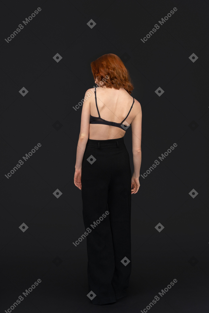 A backside view of the sexy young woman standing on the black background posing and looking to the left
