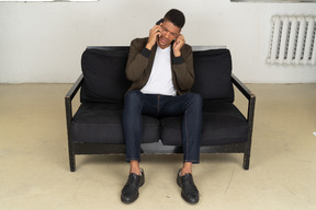 Front view of a perplexed young man sitting on a sofa and talking on his phone