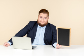 坐在膝上型计算机前面和拿着小黑板的年轻超重人