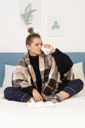 Vue de face d'une jeune femme malade en pyjama enveloppée dans une couverture à carreaux au lit en se mouchant