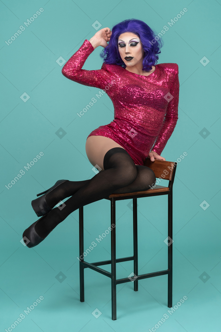 Drag queen sentado em um banquinho com a mão levantada até a cabeça