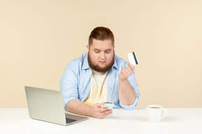 Young overweight man sitting at the table, using phone and doing online shopping