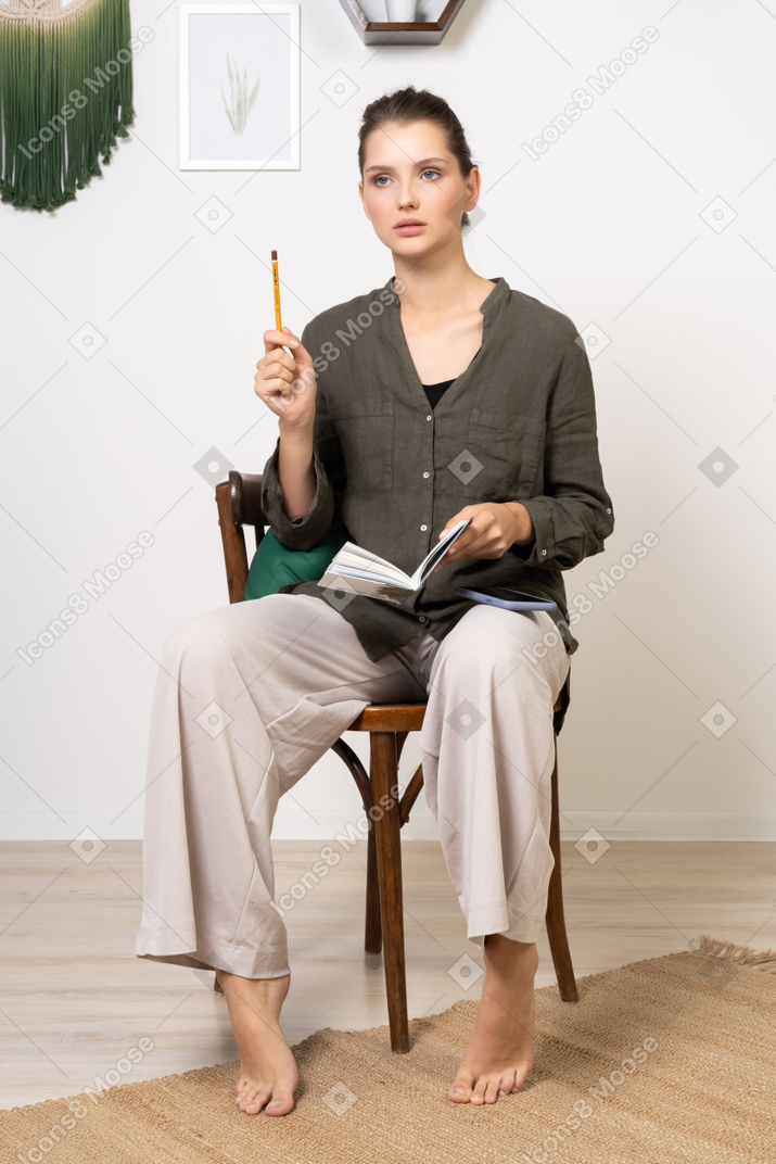 Vista frontale di una giovane donna premurosa che indossa abiti da casa seduta su una sedia con matita e taccuino