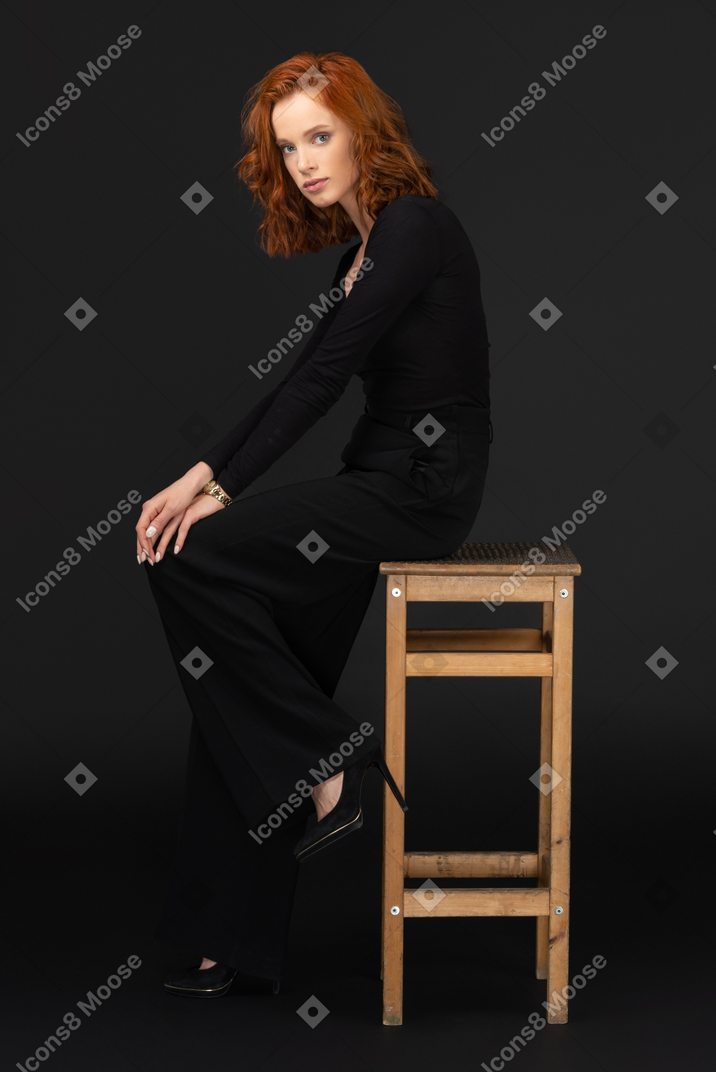 A side view of the sexy woman sitting on the tall wooden chair and looking to the camera