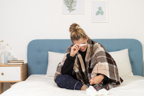Vista frontal de uma jovem sorridente doente de pijama, enrolada em um cobertor xadrez, verificando sua temperatura