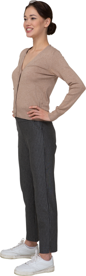 Vista di tre quarti di una donna sorridente in pullover e pantaloni che mette le mani sui fianchi