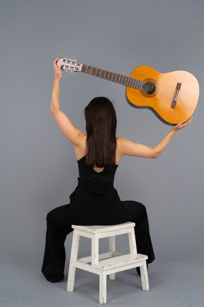 Back view of a young lady in black suit holding the guitar over head and sitting on stool