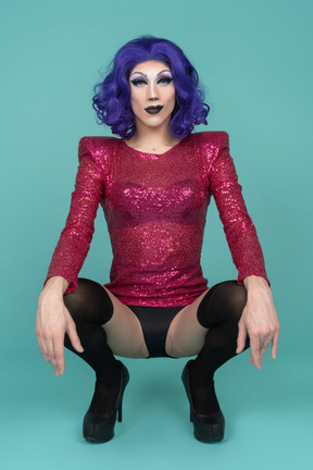Retrato de uma drag queen em vestido de lantejoulas rosa de cócoras com as mãos apoiadas nos joelhos
