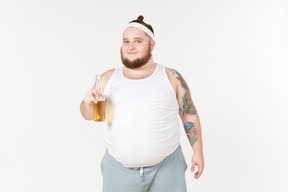 Um homem gordo no sportswear segurando uma garrafa de cerveja e olhando satisfeito