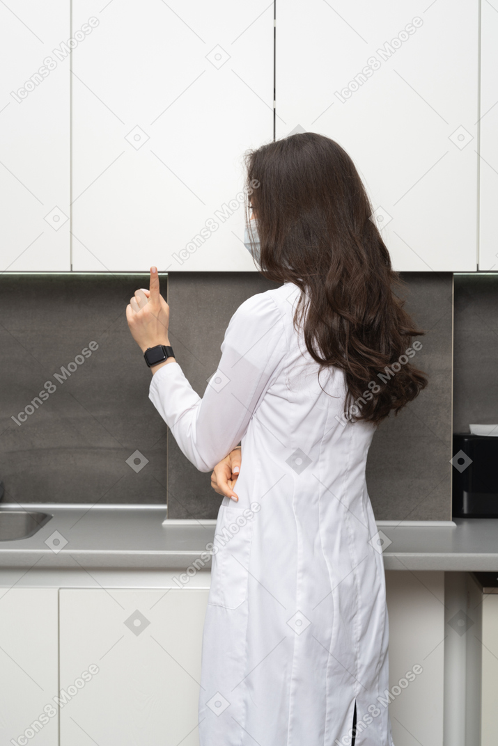 Vista de três quartos das costas de uma médica usando máscara apontando o dedo