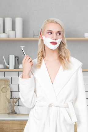 A woman in a white robe is shaving her face with a razor