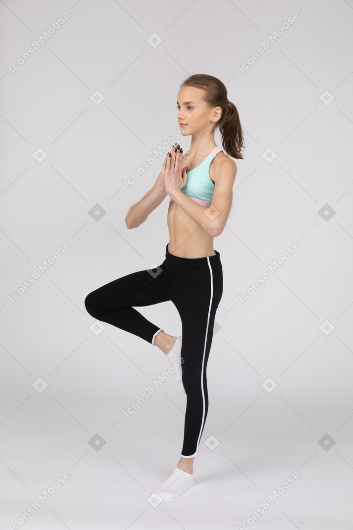 Vista de três quartos de uma adolescente em roupas esportivas, se equilibrando em uma perna e de mãos dadas
