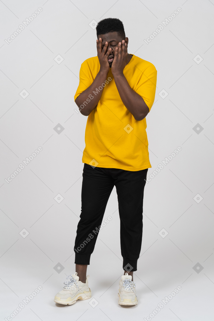 Front view of an exhausted young dark-skinned man in yellow t-shirt touching his face