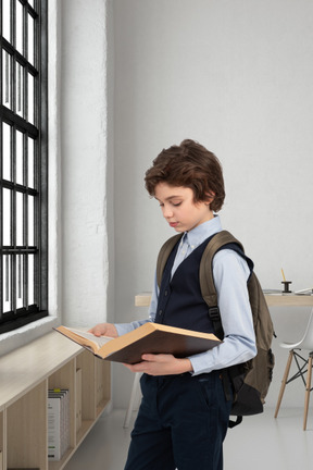 A boy with a backpack is reading a book in front of a window