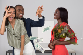 Woman doing ok sign and another woman holding bouquet while man behind them throwing dollar bills