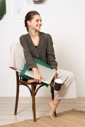 Vorderansicht einer lächelnden jungen frau, die auf einem stuhl sitzt und ihren laptop und ihre kaffeetasse hält