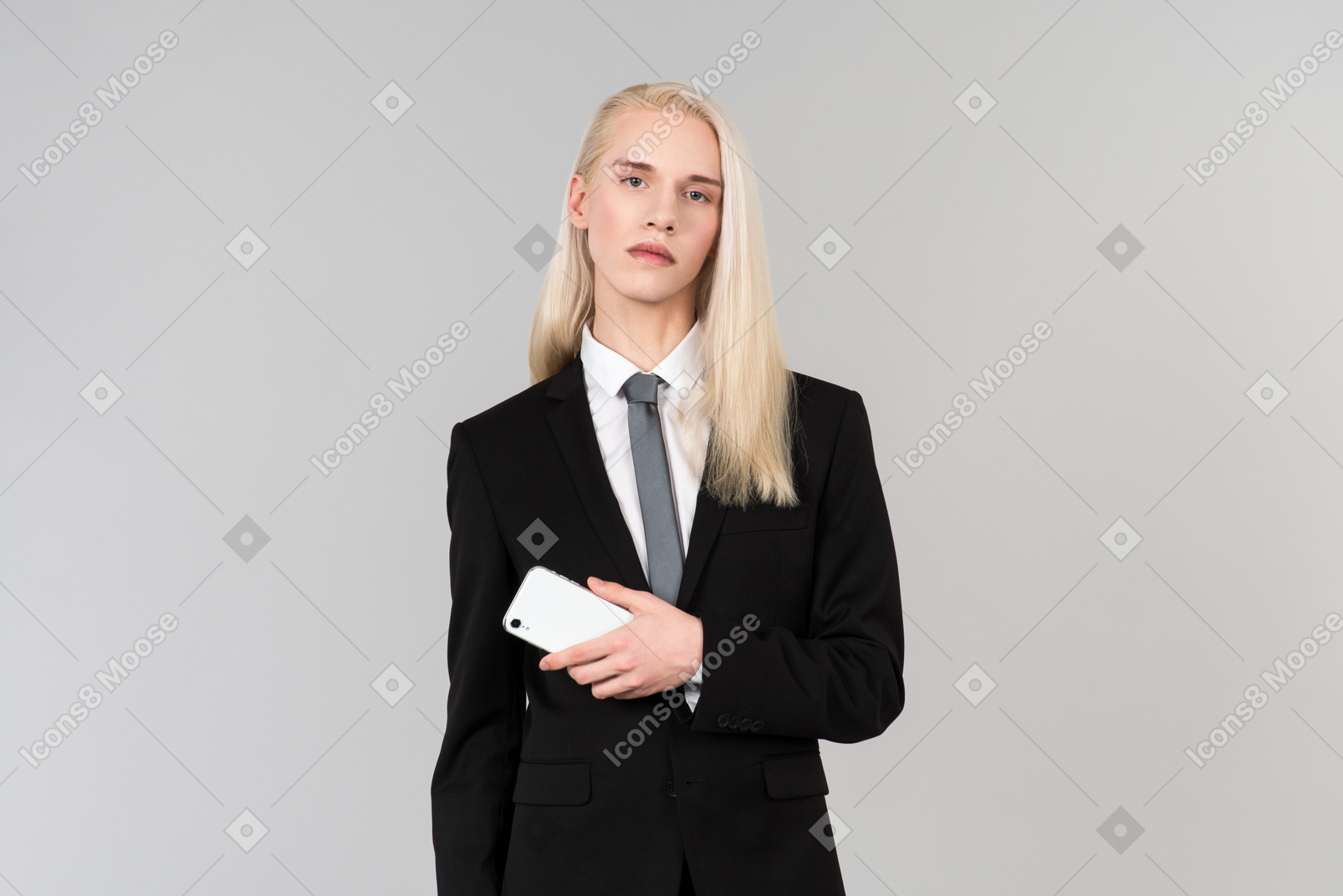Jeune bel homme aux longs cheveux blonds, vêtu d'un costume noir et d'une cravate, debout contre le fond gris uni