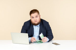 Молодой офисный работник с избыточным весом сидит за столом и считает деньги