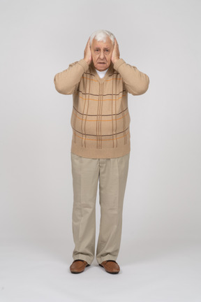 Vista frontal de um velho assustado em roupas casuais, cobrindo os ouvidos com as mãos e olhando para a câmera