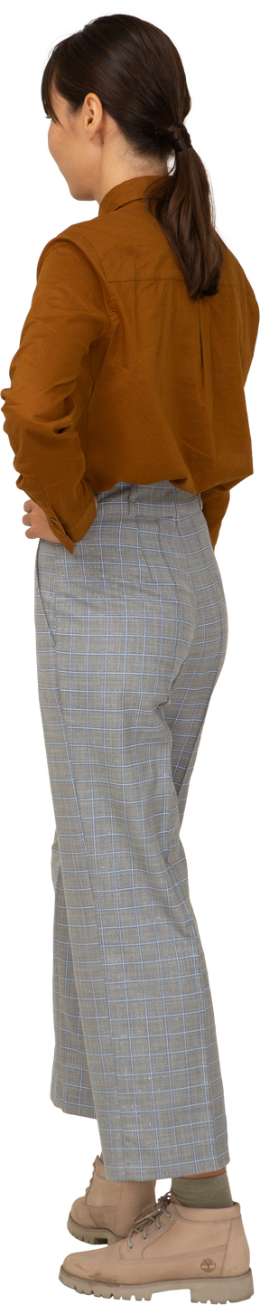 Vista traseira de três quartos de uma jovem mulher asiática fazendo beicinho de calça e blusa colocando as mãos nos quadris