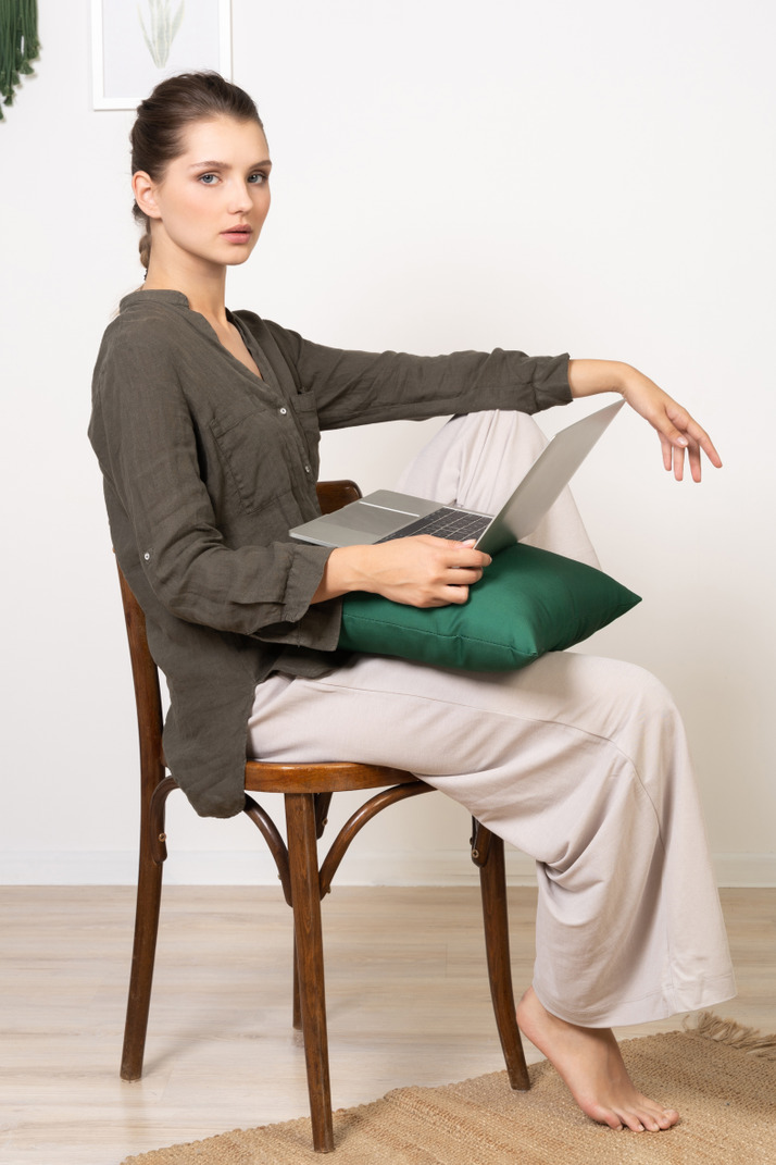 Side view of a young woman wearing home clothes sitting on a chair with a laptop
