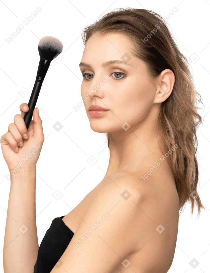 Вид сбоку чувственной молодой женщины, держащей кисть для макияжа и смотрящей в камеру