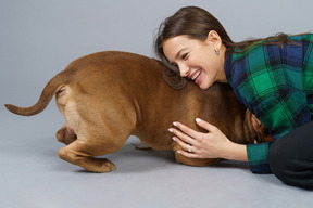 Foto lateral de uma jovem mulher de camisa xadrez abraçando um bulldog marrom