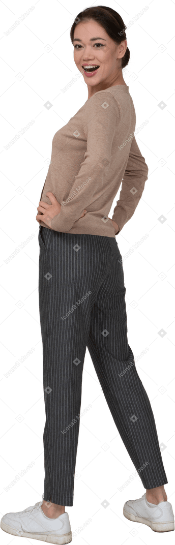 Vista posteriore di tre quarti di una giovane donna sorridente in pullover e pantaloni mettendo le mani sui fianchi