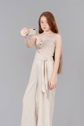 Teenage girl dressed in beige overalls making a selfie with smartphone