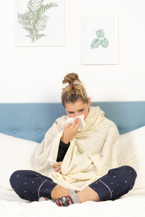 Front view of a young lady wrapped in white blanket sitting in bed and blowing nose