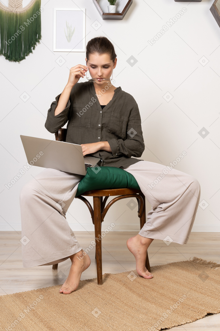 Вид спереди молодой женщины в домашней одежде, сидящей на стуле с ноутбуком и надевающей очки