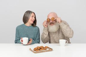 Elder man covering his eyes with cookies sitting next to his wife