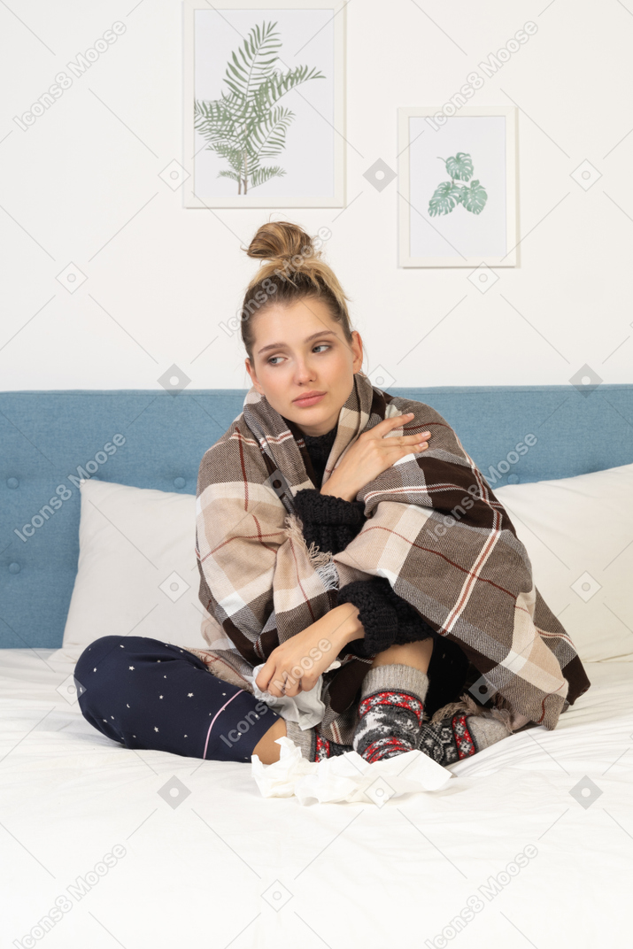 Front view of a sad ill young lady in pajamas wrapped in checked blanket in bed
