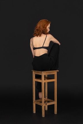 A backside view of the beautiful woman dressed in black pants and bra, sitting on the wooden chair and holding her hand on the leg
