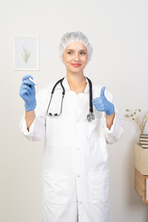 Front view of a young female doctor with stethoscope holding thermometer and showing thumb up