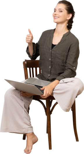 Front view of a smiling young woman sitting on a chair with a laptop & showing thumb up