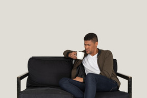 Front view of a young grimacing man sitting on a sofa with a cup of coffee