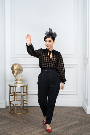 Stylish young woman raising hand in greeting while stepping forward