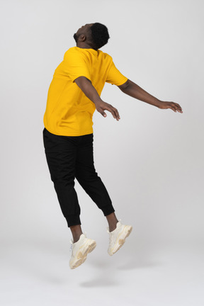 Vista das costas de três quartos de um homem jovem de pele escura pulando em uma camiseta amarela estendendo as mãos