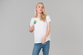 Apuesto joven con largo cabello rubio, de pie contra el fondo gris, vestido con jeans azules y una camiseta blanca con la insignia lgbt