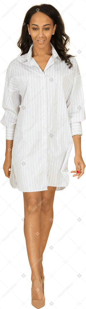 Vue de face d'une jeune femme à la peau foncée marche souriante en robe blanche