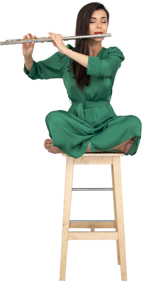 Девушка в полный рост, играющая на кларнете, сидящая со скрещенными ногами на деревянном стуле