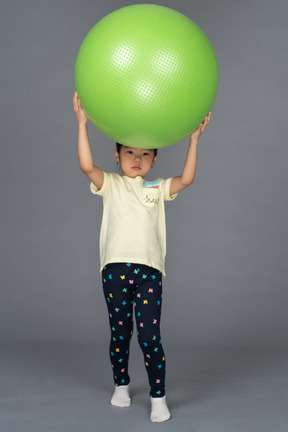 Маленькая девочка держит над головой зеленый фитбол