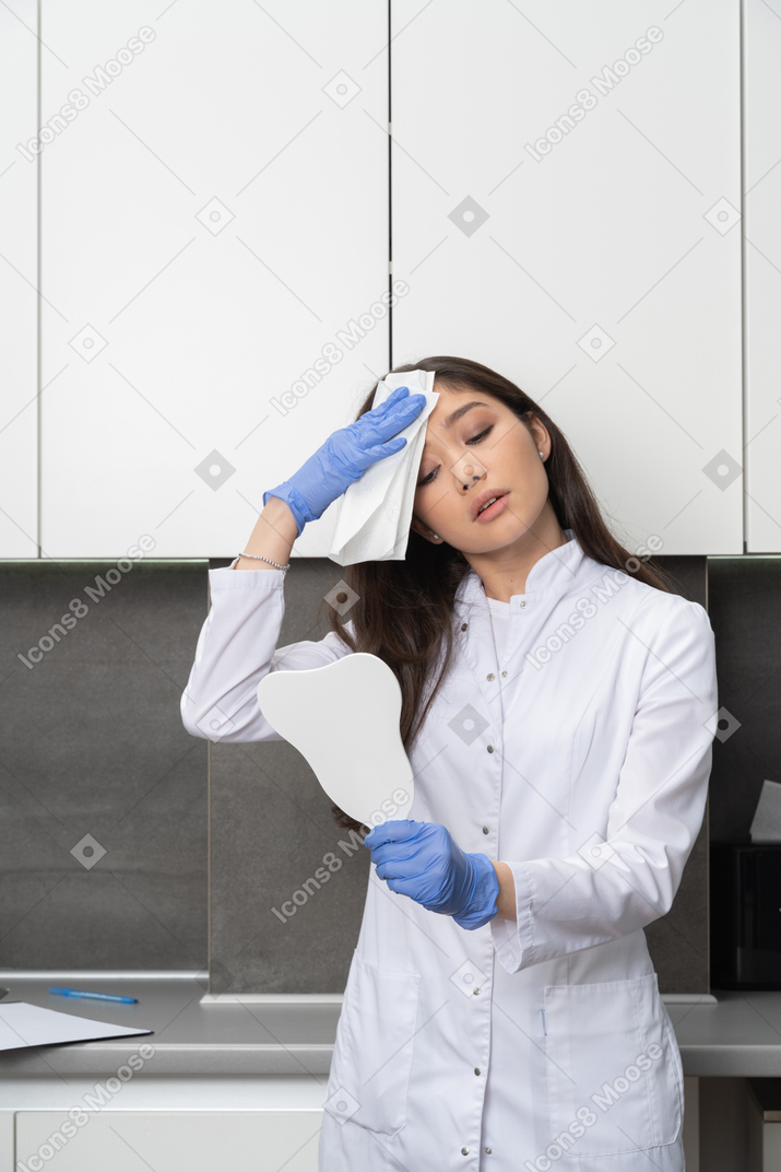Vista frontale di una dottoressa stanca guardarsi allo specchio e asciugandosi la fronte