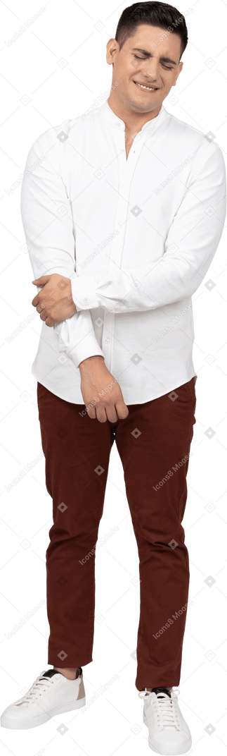 Vista frontal de um jovem latino segurando seu braço e fechando os olhos em desgosto com um sorriso